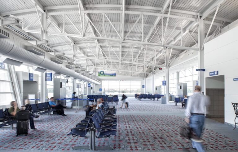 Airport entrance hallway with people walking and sitting on blue chairs.